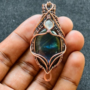 Peut inclure: Pendentif artisanal en fil de cuivre avec une pierre précieuse ovale et irisée. La pierre est d'un bleu-vert profond avec des reflets bleus et verts. Une petite pierre transparente est placée au-dessus. Le pendentif mesure environ 5 cm de long.