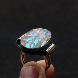 Op de afbeelding: Een ring met een ovaalvormige abalone schelp inlay met iriserende blauwe, groene en paarse tinten. De schelp is gezet in een donkere metalen zetting, geflankeerd door twee gebogen metalen zijaccenten en ondersteund door een koperkleurige metalen ringband.