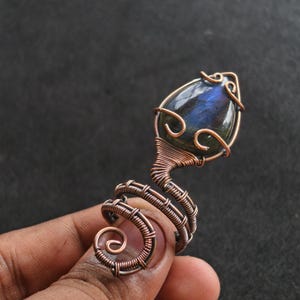May include: A copper wire-wrapped ring featuring a teardrop-shaped, iridescent blue-green gemstone. The ring's design includes a spiral and decorative swirls, showcasing the intricate wirework and the stone's natural beauty.