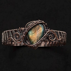 Puede incluir: Una pulsera artesanal de alambre de cobre con una piedra de labradorita en forma de corazón. La piedra muestra colores iridiscentes de azul, verde y dorado. El trabajo de alambre está intrincadamente tejido, con espirales decorativas en los lados, creando una joya única.