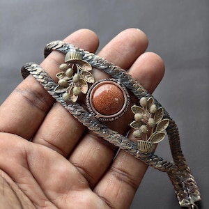 May include: A cuff bracelet featuring a textured, braided silver-tone metal band. The center is adorned with a round, shimmering copper-colored sandstone cabochon flanked by two decorative metal sprigs of leaves and small round berries.