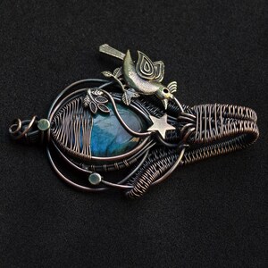 Puede incluir: Colgante artesanal con una piedra preciosa azul, alambre envuelto en tonos cobre y plata. Un peque&ntilde;o p&aacute;jaro y una estrella acent&uacute;an el dise&ntilde;o. El colgante mide aproximadamente 5 cm de largo y 4 cm de ancho.