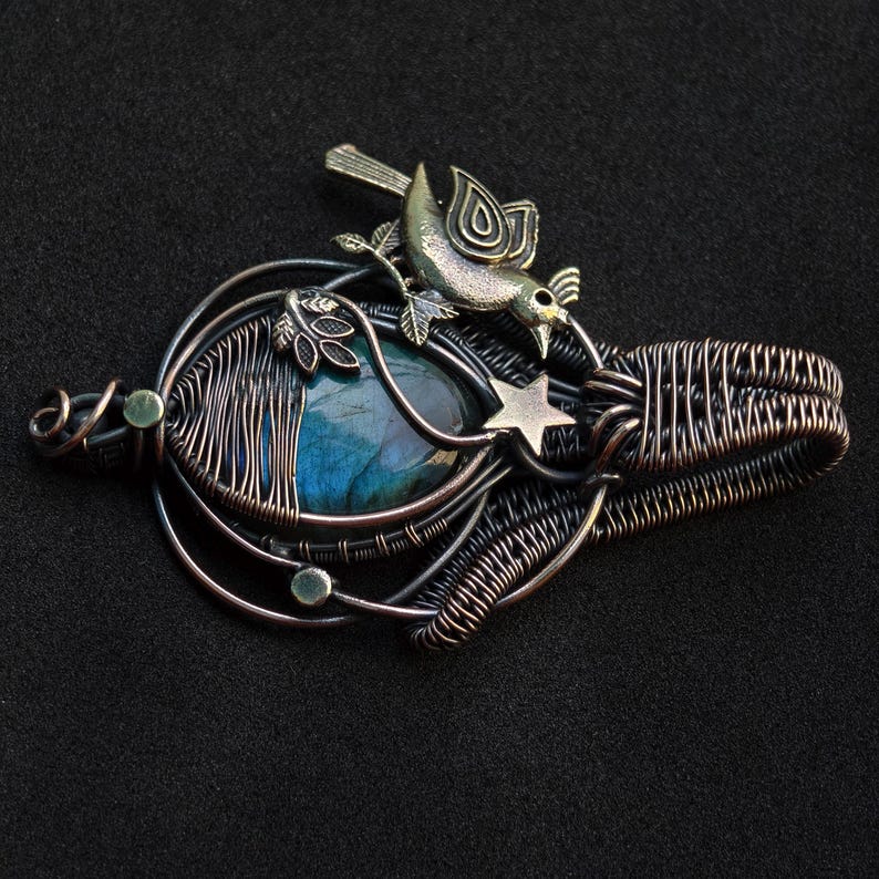 Puede incluir: Colgante artesanal con una piedra preciosa azul, alambre envuelto en tonos cobre y plata. Un peque&ntilde;o p&aacute;jaro y una estrella acent&uacute;an el dise&ntilde;o. El colgante mide aproximadamente 5 cm de largo y 4 cm de ancho.