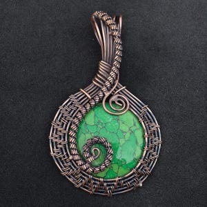 May include: Handmade pendant featuring a round, vibrant green stone with a unique crackled pattern. The stone is encased in intricate copper wire, with a woven design around the stone and a decorative spiral at the top. The pendant is approximately 2 inches long.