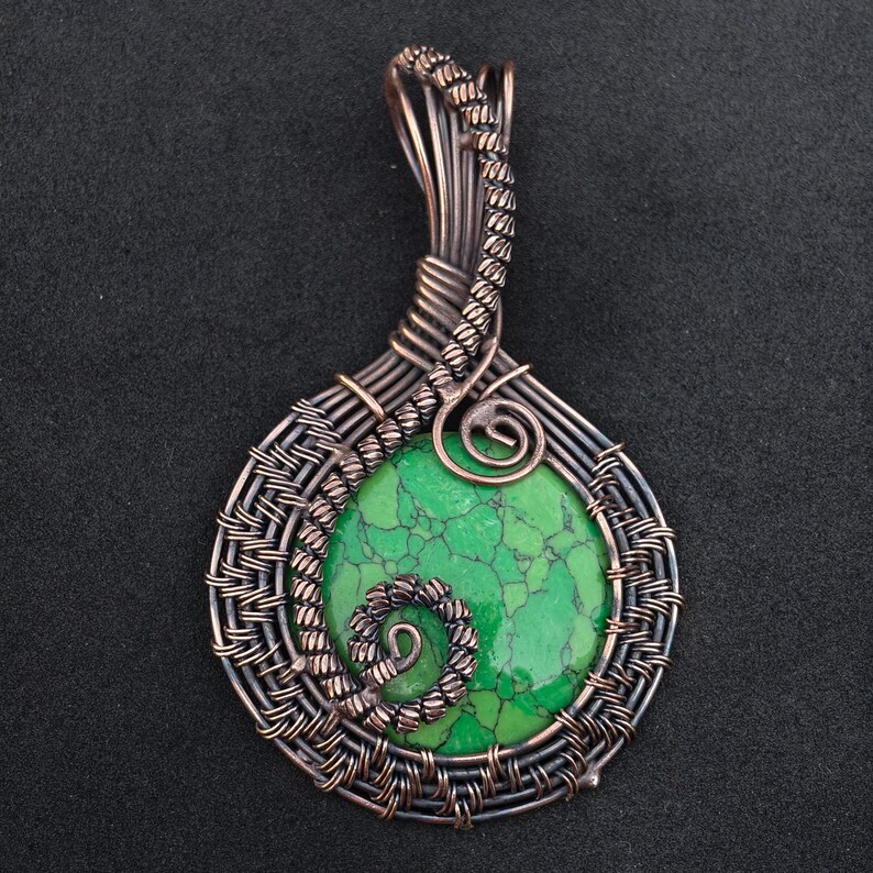 May include: Handmade pendant featuring a round, vibrant green stone with a unique crackled pattern. The stone is encased in intricate copper wire, with a woven design around the stone and a decorative spiral at the top. The pendant is approximately 2 inches long.