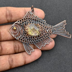 Peut inclure: Pendentif poisson en cuivre fait à la main avec une pierre précieuse en son centre. Le poisson présente des détails complexes en fil de fer, notamment les nageoires, la queue et les écailles. La pierre précieuse est de couleur jaune doré clair. Le pendentif est tenu dans une main sur fond sombre.