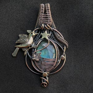 Puede incluir: Colgante artesanal con dise&ntilde;o de alambre. Presenta una piedra ovalada con colores azules y verdes. Un peque&ntilde;o p&aacute;jaro y una estrella adornan el colgante, hecho de alambre de cobre y plateado.