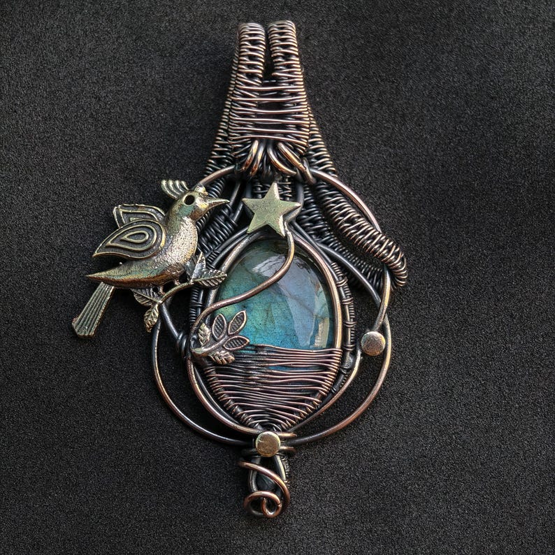 Puede incluir: Colgante artesanal con dise&ntilde;o de alambre. Presenta una piedra ovalada con colores azules y verdes. Un peque&ntilde;o p&aacute;jaro y una estrella adornan el colgante, hecho de alambre de cobre y plateado.