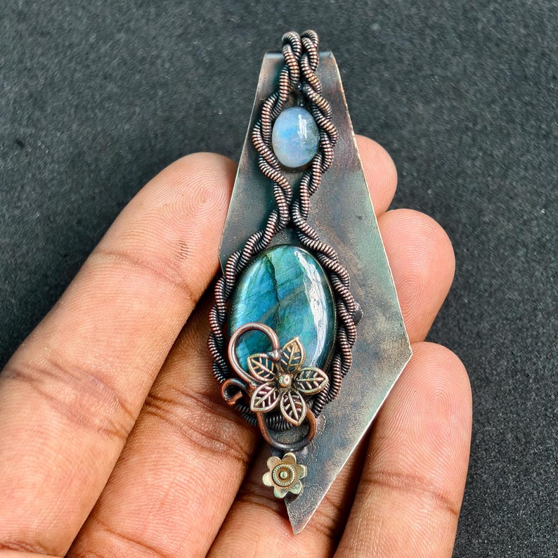 May include: A handcrafted pendant featuring a large, oval labradorite stone with blue and green hues, accented by a smaller moonstone. The stones are set in a copper wire wrap design with a floral embellishment. The pendant is approximately 7.6 cm long.