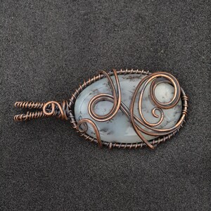 Peut inclure: Pendentif ovale avec une pierre p&acirc;le vein&eacute;e de gris, envelopp&eacute;e de fil de cuivre. Le fil de fer comprend des volutes d&eacute;coratives et une b&eacute;li&egrave;re torsad&eacute;e. Le pendentif est pos&eacute; sur un fond gris fonc&eacute;. Bijou unique.