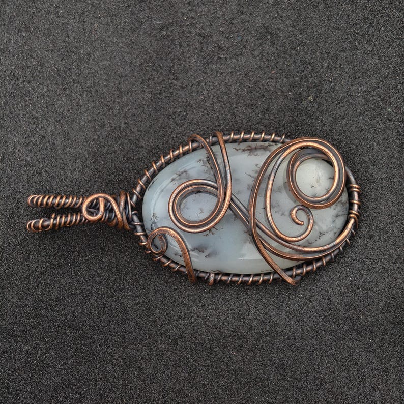 Peut inclure: Pendentif ovale avec une pierre p&acirc;le vein&eacute;e de gris, envelopp&eacute;e de fil de cuivre. Le fil de fer comprend des volutes d&eacute;coratives et une b&eacute;li&egrave;re torsad&eacute;e. Le pendentif est pos&eacute; sur un fond gris fonc&eacute;. Bijou unique.