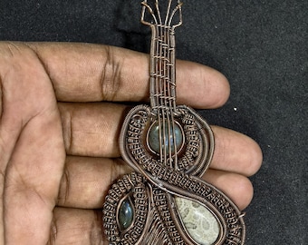 May include: A copper wire pendant in the shape of a guitar, featuring a detailed wire wrap design. The pendant incorporates three stones: a round, blue-green stone, a teardrop-shaped stone, and a small, oval stone. The guitar-shaped pendant is held in a hand.