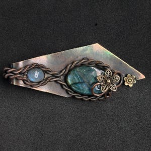 May include: Handmade copper-toned metal barrette. It features an oval labradorite stone and a smaller, round, light blue stone. The barrette has a floral design and a small flower accent.