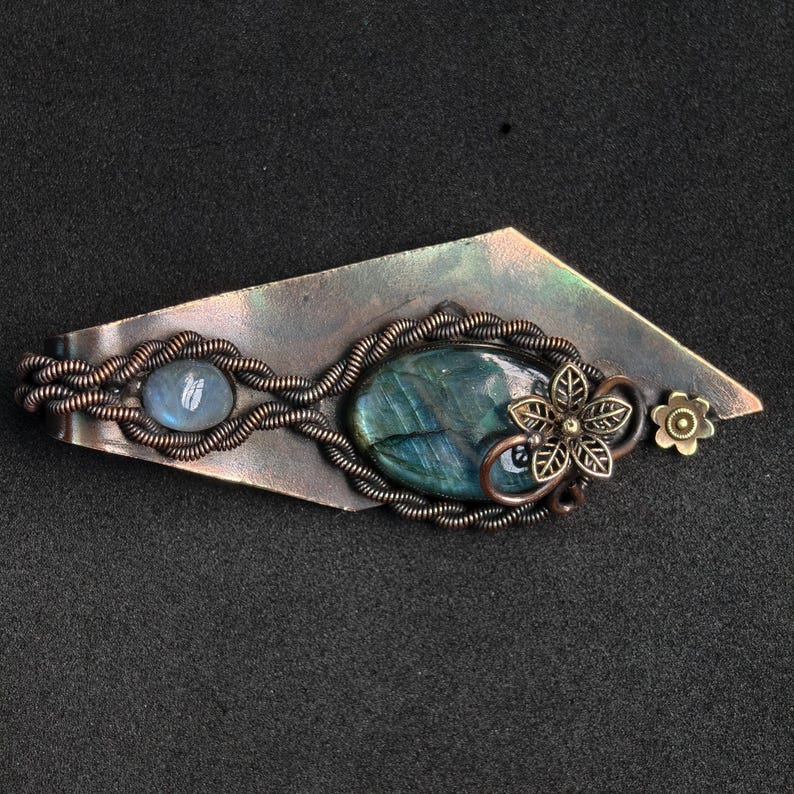 May include: Handmade copper-toned metal barrette. It features an oval labradorite stone and a smaller, round, light blue stone. The barrette has a floral design and a small flower accent.
