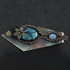 May include: A handcrafted copper-toned metal barrette with a large, oval, blue-green labradorite stone. The barrette features a flower and a smaller, round, blue moonstone. The design incorporates wire wrapping and floral accents.