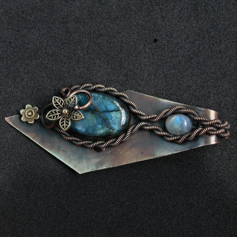 May include: A handcrafted copper-toned metal barrette with a large, oval, blue-green labradorite stone. The barrette features a flower and a smaller, round, blue moonstone. The design incorporates wire wrapping and floral accents.