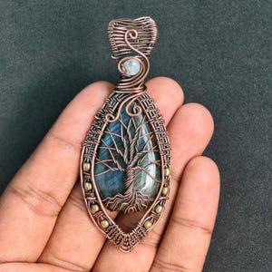 May include: A handcrafted copper pendant featuring a tree of life design. The pendant has a teardrop shape with a blue-green stone center. The tree's branches and roots are intricately detailed. Accented with small gold-colored beads and a light blue stone.