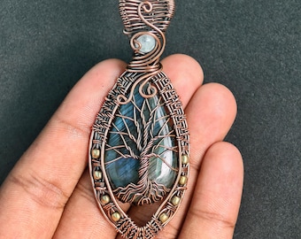 Peut inclure: Pendentif en cuivre fait à la main avec un motif d'arbre de vie. Le pendentif a une forme de goutte d'eau avec un centre en pierre bleu-vert. Les branches et les racines de l'arbre sont finement détaillées. Orné de petites perles dorées et d'une pierre bleu clair.