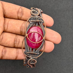 May include: A handcrafted wire-wrapped bracelet featuring a large, oval, deep red gemstone. The bracelet's intricate design incorporates copper and silver-toned wire, creating a unique and artistic piece of jewellery.
