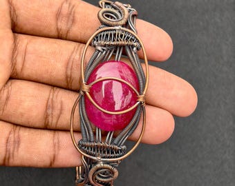 May include: A handcrafted wire-wrapped bracelet featuring a large, oval, deep red gemstone. The bracelet's intricate design incorporates copper and silver-toned wire, creating a unique and artistic piece of jewellery.