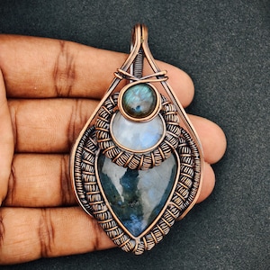May include: A handcrafted copper pendant featuring a teardrop-shaped labradorite stone, a round moonstone, and a small round labradorite. The pendant is intricately wire-wrapped with a detailed design, showcasing the natural beauty of the gemstones.