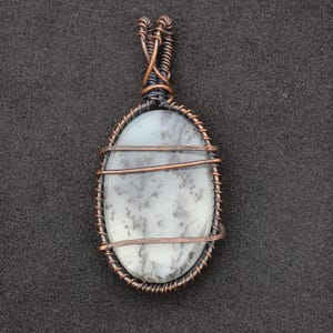 Peut inclure: Pendentif ovale avec une pierre blanche vein&eacute;e de gris, sertie dans du fil de cuivre. Le fil est soigneusement enroul&eacute; autour de la pierre, formant un cadre d&eacute;coratif et une b&eacute;li&egrave;re pour un collier. Le pendentif est pr&eacute;sent&eacute; sur fond sombre.