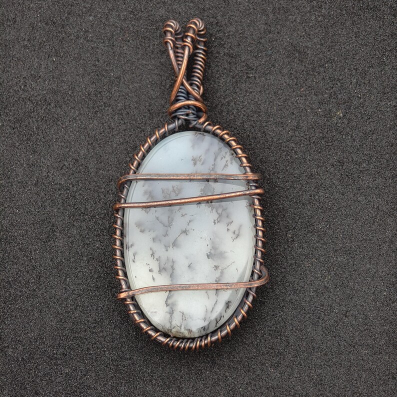 Peut inclure: Pendentif ovale avec une pierre blanche vein&eacute;e de gris, sertie dans du fil de cuivre. Le fil est soigneusement enroul&eacute; autour de la pierre, formant un cadre d&eacute;coratif et une b&eacute;li&egrave;re pour un collier. Le pendentif est pr&eacute;sent&eacute; sur fond sombre.
