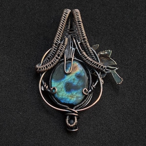 Puede incluir: Colgante artesanal con una piedra ovalada de labradorita con tonos iridiscentes azules y verdes, engastada en intrincados alambres de cobre. Un peque&ntilde;o dise&ntilde;o estilizado de p&aacute;jaro y hoja est&aacute; adjunto, agregando un elemento natural a la joya.