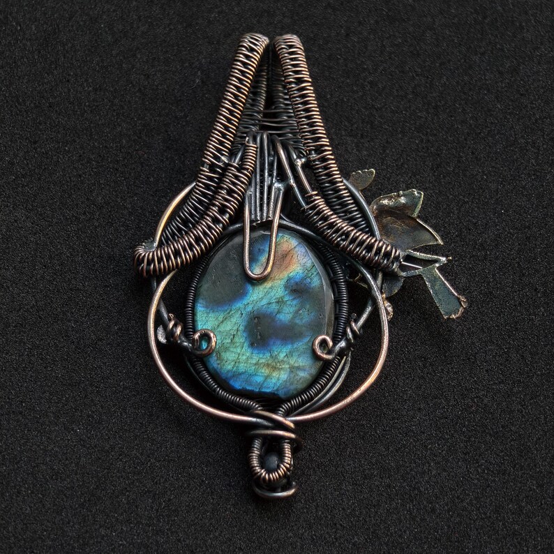 Puede incluir: Colgante artesanal con una piedra ovalada de labradorita con tonos iridiscentes azules y verdes, engastada en intrincados alambres de cobre. Un peque&ntilde;o dise&ntilde;o estilizado de p&aacute;jaro y hoja est&aacute; adjunto, agregando un elemento natural a la joya.