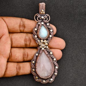 May include: A wire-wrapped pendant featuring a teardrop-shaped light blue moonstone at the top and a larger teardrop-shaped pale pink rose quartz at the bottom. The stones are encased in intricate copper wire coils with small metal bead accents.