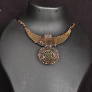 May include: A handcrafted necklace with a bronze-coloured crescent-shaped upper section and a circular pendant. The pendant features a central green stone surrounded by a wire-wrapped design. The necklace is displayed on a black mannequin.