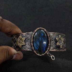 May include: A cuff bracelet featuring a central oval labradorite gemstone with blue iridescence, encased in copper wire wrapping. The band has a dark, braided texture with two brass-colored flower accents on either side and a small metal chain link.