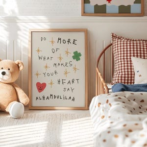 Puede incluir: Una impresión artística enmarcada con el texto "DO MORE OF WHAT MAKES YOUR HEART SAY ALHAMDULILAH" en negro, con ilustraciones de estrellas y tréboles. Un oso de peluche y una cama con una almohada a cuadros están en el fondo.