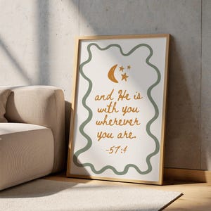 Puede incluir: Un cuadro enmarcado con la cita "and He is with you wherever you are". El texto es en un tono cálido y terroso, con un gráfico de luna creciente y estrellas. El marco es de madera clara, con un borde verde ondulado.