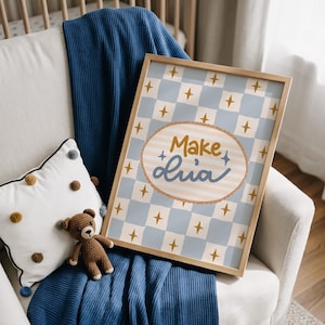 May include: A framed art print with a light blue and white checkered pattern and gold star accents. The print features the text "Make + Lia" in a whimsical font. The artwork is displayed on a white armchair with a blue blanket and a teddy bear.