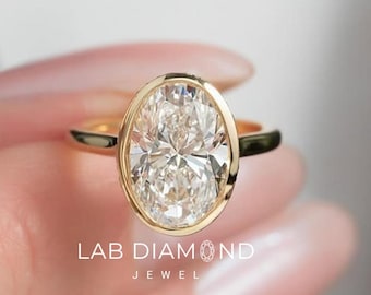May include: A gold ring featuring a large, oval-cut diamond. The diamond is set in a bezel setting, and the band is a simple, polished gold. The ring is held up by a hand, with the text "LAB DIAMOND JEWEL" visible.