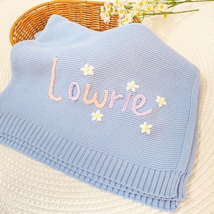 May include: A pale blue knitted baby blanket embroidered with the name "Lowrie" in pink and lilac. The blanket features small white and yellow flower appliqués. The blanket is folded and placed in a wicker basket.