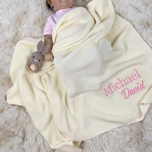 May include: A cream knitted baby blanket with the name "Michael David" embroidered in pink. A small, crocheted bunny rattle with a wooden ring is also visible. The blanket is on a fluffy, cream-coloured surface.