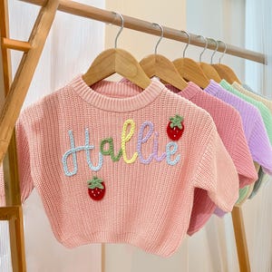 Puede incluir: Una fila de suéteres pastel para niños colgados en perchas de madera. El suéter delantero es de color rosa claro con el nombre "Hallie" bordado en hilo colorido, junto con dos aplicaciones de fresas. Otros suéteres son rosas, morados, verdes y amarillos.