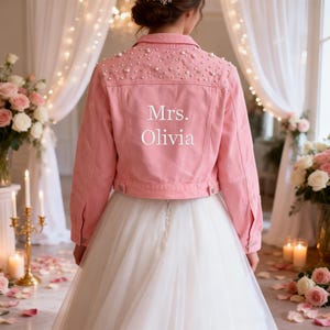 Puede incluir: Una chaqueta vaquera de color rosa claro con adornos de perlas blancas en la parte superior de la espalda y los hombros. La parte posterior de la chaqueta muestra el texto impreso en blanco "Mrs. Olivia" centrado bajo las perlas.
