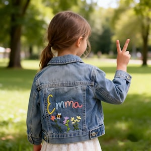 May include: A light blue denim jacket featuring the embroidered name Emma in colourful yarn on the back. Below the name, there is an embroidered design of a small bee flying toward three stems of colourful flowers with green leaves and petals.