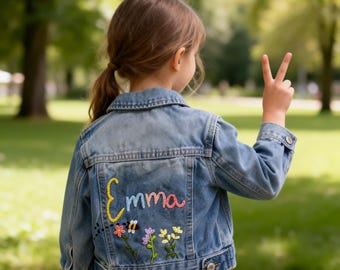 May include: A light blue denim jacket featuring the embroidered name Emma in colourful yarn on the back. Below the name, there is an embroidered design of a small bee flying toward three stems of colourful flowers with green leaves and petals.