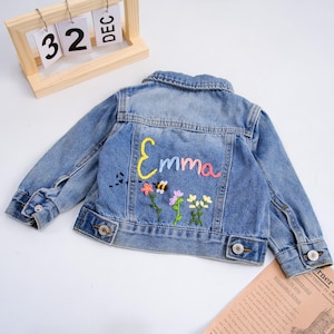 May include: A light blue denim jacket featuring a fold-down collar, long sleeves with buttoned cuffs, and two chest pockets. The back is embroidered with the name Emma in colourful thread, accompanied by a small bee and three floral stems.