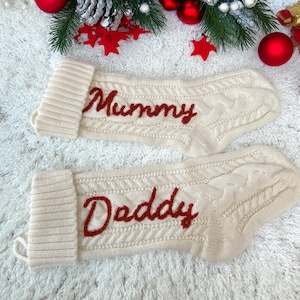 Może przedstawiać: Dwie kremowe skarpety świąteczne z wyhaftowanymi na czerwono słowami "Mummy" i "Daddy". Skarpety mają wzór warkocza i są ozdobione ozdobami świątecznymi.