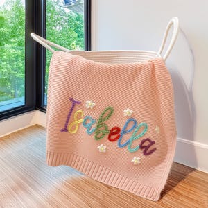 May include: A pale pink knitted blanket embroidered with the name "Isabella" in various colours. The blanket is draped over a white rope basket. Small white flower details are also embroidered on the blanket.