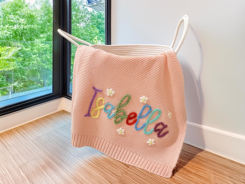 May include: A pale pink knitted blanket embroidered with the name "Isabella" in various colours. The blanket is draped over a white rope basket. Small white flower details are also embroidered on the blanket.
