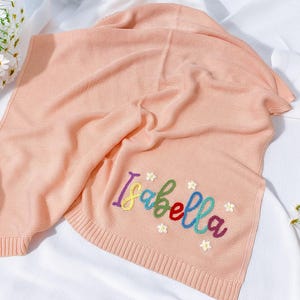 May include: A light peach knitted blanket featuring the name "Isabella" embroidered in rainbow colours. The blanket has a ribbed texture and is decorated with small embroidered flowers. The blanket is folded on a white surface.