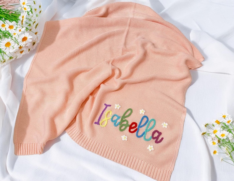 May include: A light peach knitted blanket featuring the name "Isabella" embroidered in rainbow colours. The blanket has a ribbed texture and is decorated with small embroidered flowers. The blanket is folded on a white surface.