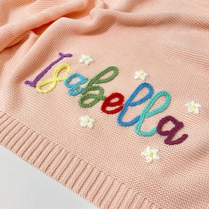 May include: A light peach knitted baby blanket featuring the name "Isabella" embroidered in colourful thread. Small white flower accents surround the name. The blanket has a ribbed border.