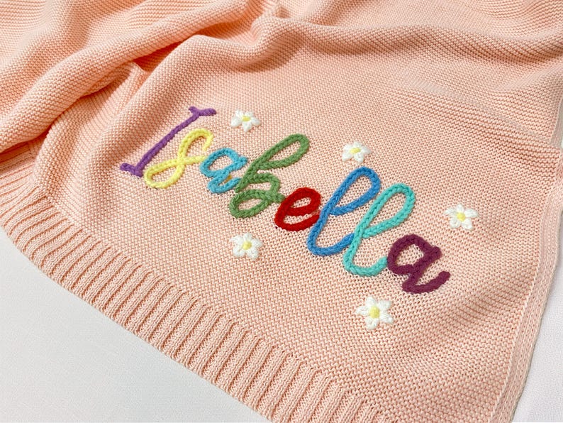 May include: A light peach knitted baby blanket featuring the name "Isabella" embroidered in colourful thread. Small white flower accents surround the name. The blanket has a ribbed border.